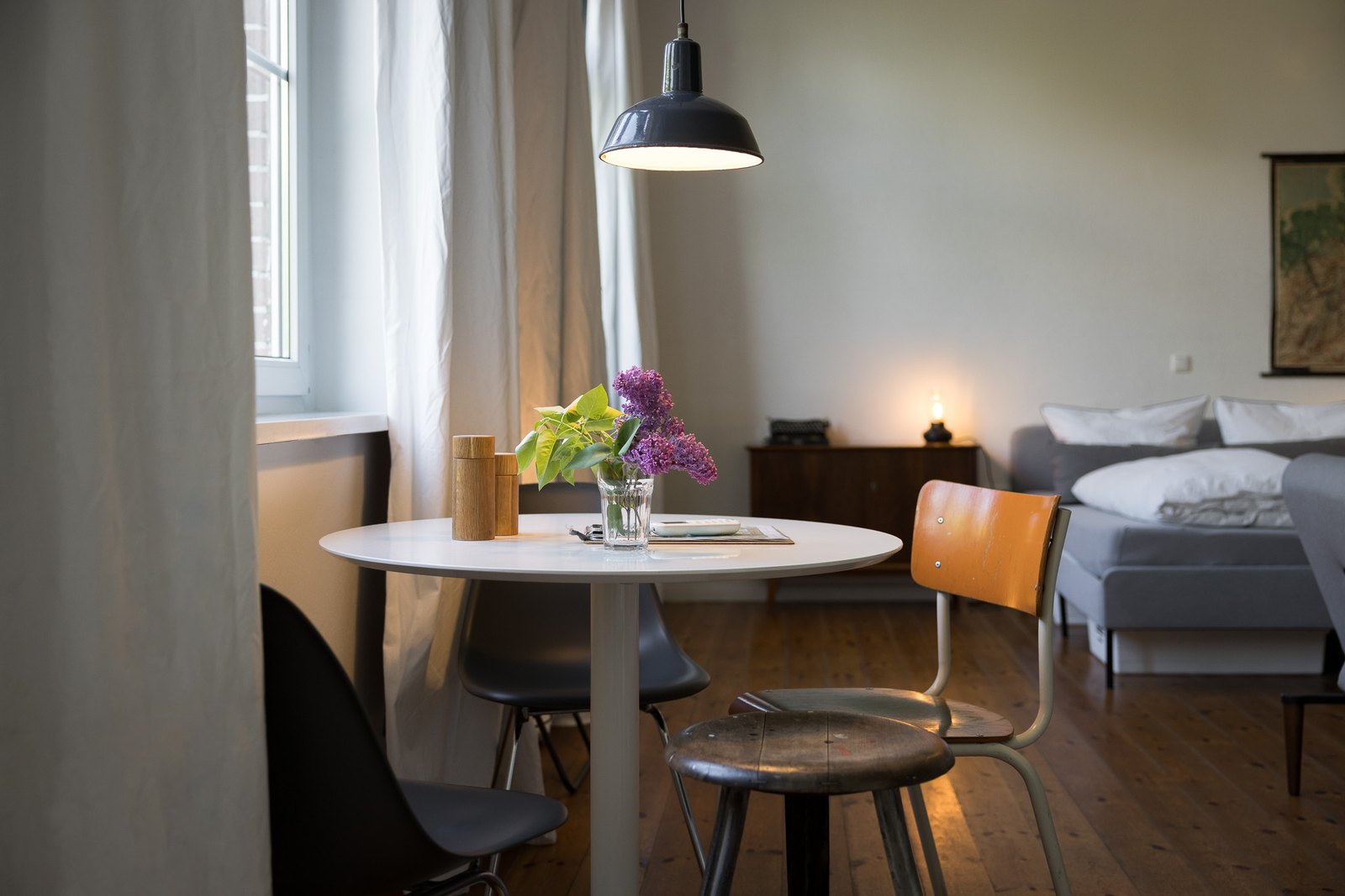 Runder Esstisch mit Flieder am Fenster, dahinter das Bett