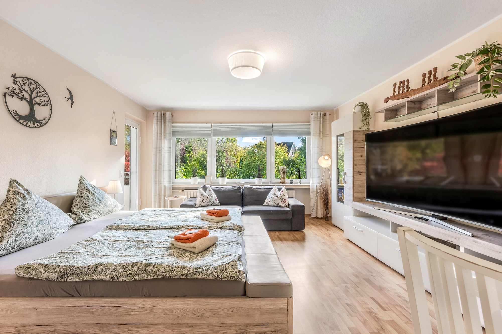 Wohn- und Schlafraum mit Doppelbett, Sofa und großem Fernseher, Fensterfront ins Grüne